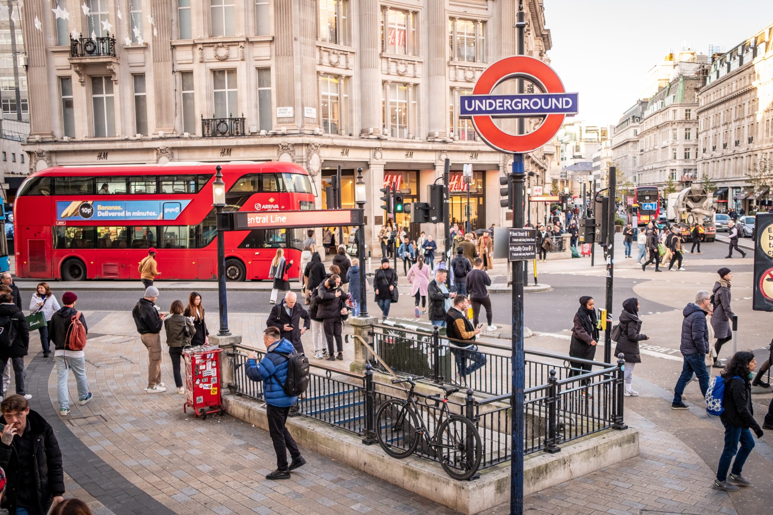 A busy London street scene with people walking, a red double-decker bus, and an Underground sign above stairs leading to a subway entrance, surrounded by historic buildings and shops.