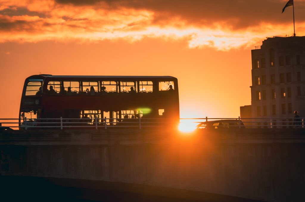 A red double-decker bus crosses a bridge at sunset, with silhouettes of passengers visible and the sun shining brightly behind the bridge.
