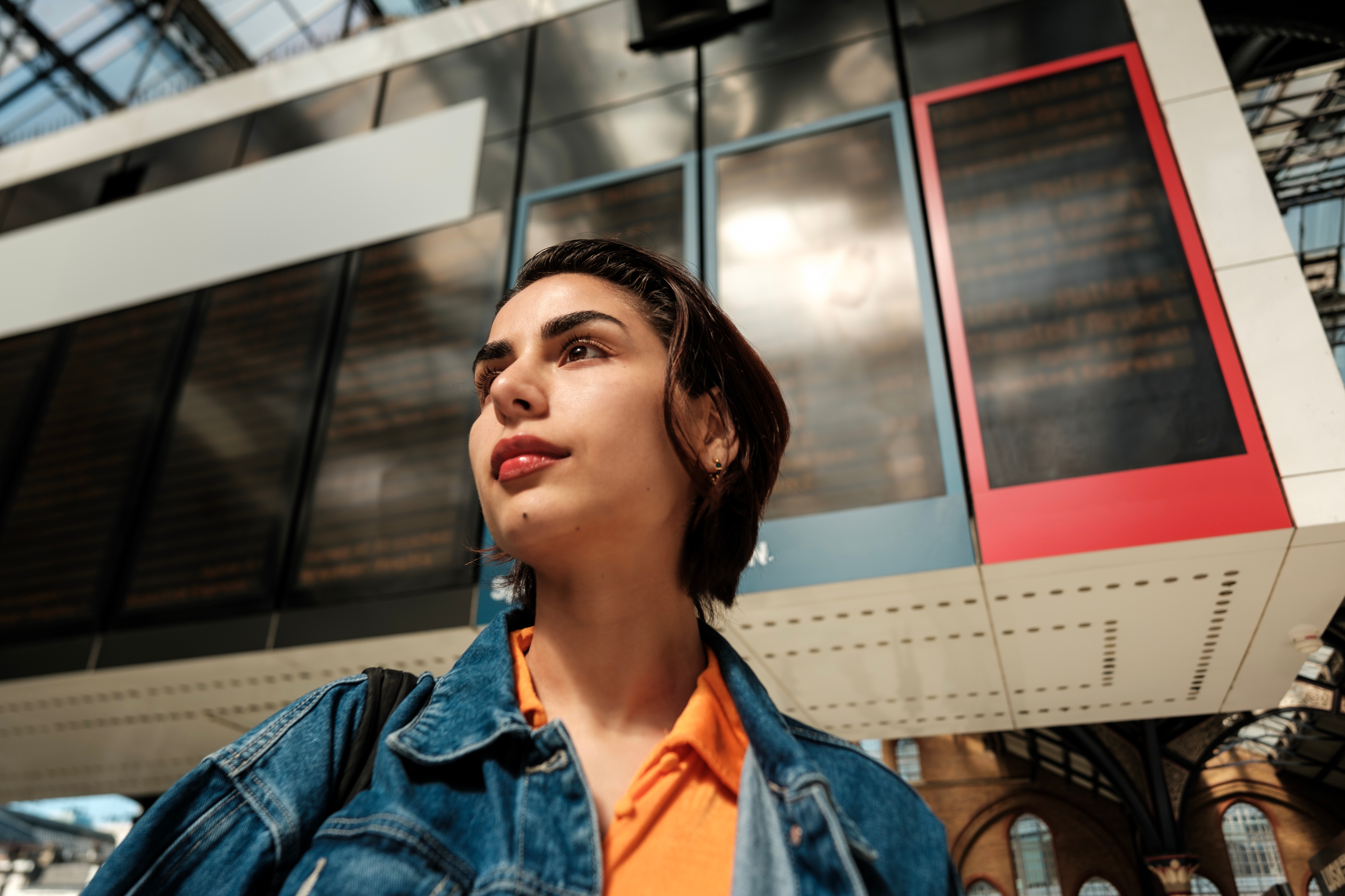 A person with short dark hair wearing a denim jacket and orange shirt stands in front of a large, blurred digital display board at a train station, looking off into the distance.