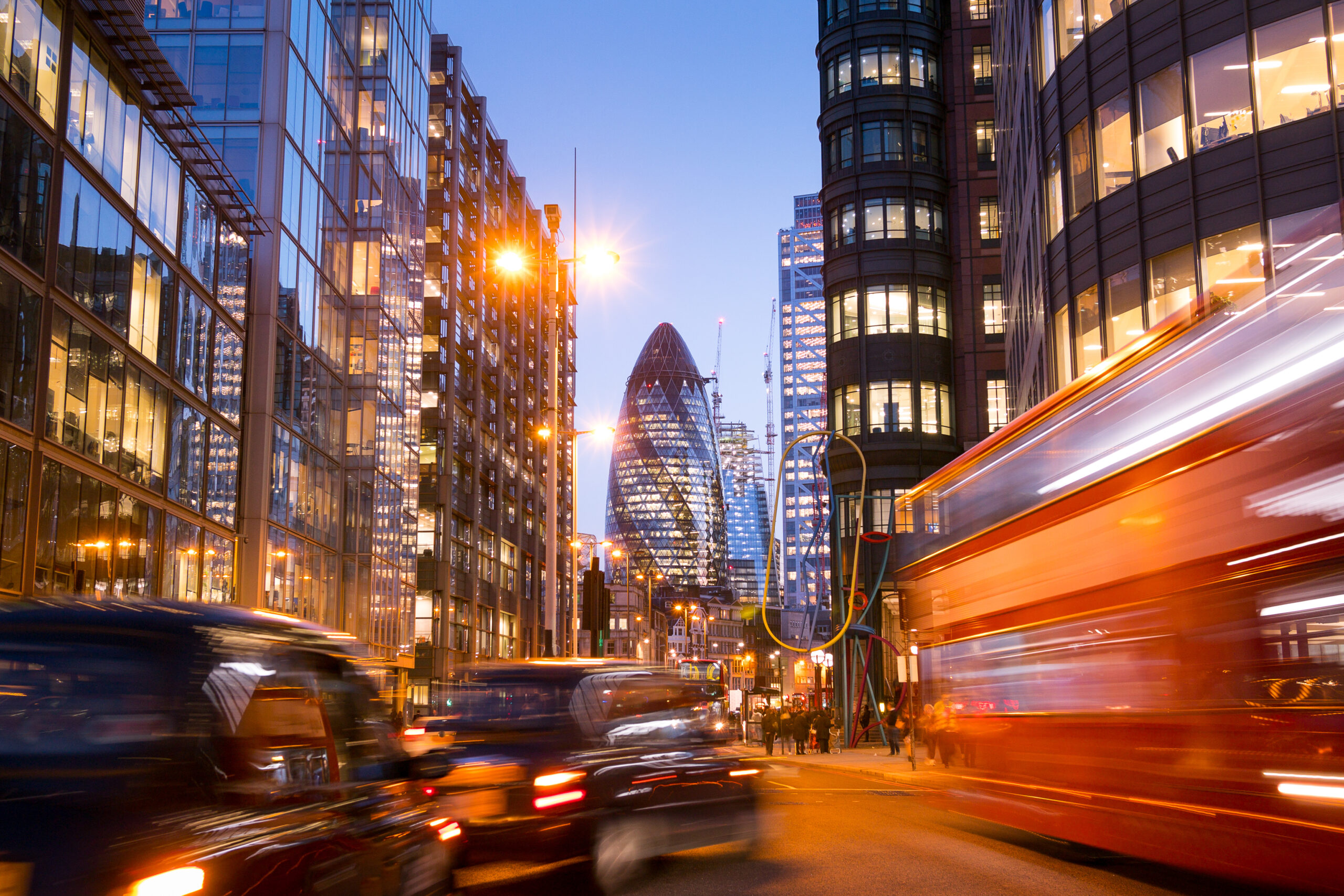 Busy London street at dusk with blurred red double-decker bus and black cabs, modern glass buildings, and the Gherkin skyscraper illuminated in the background.