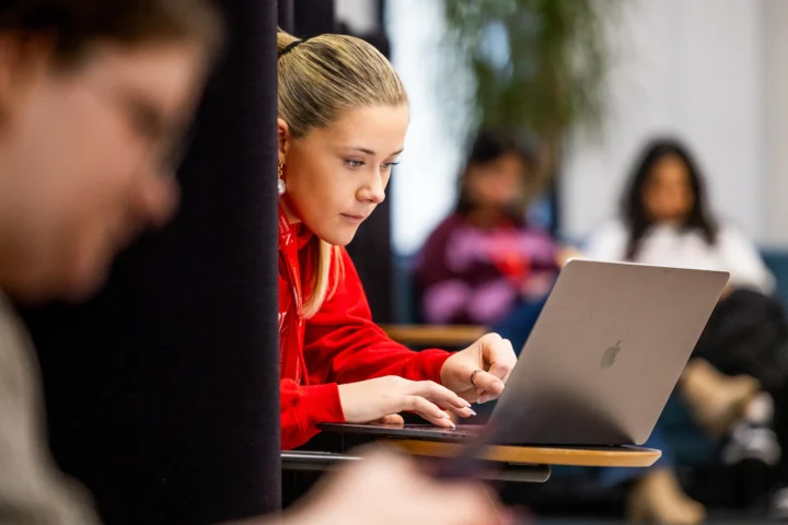 A young woman in a red hoodie focuses intently on her laptop while seated at a desk, perhaps researching how to apply through Clearing. Other people sit blurred in the background, suggesting a busy classroom or study area.