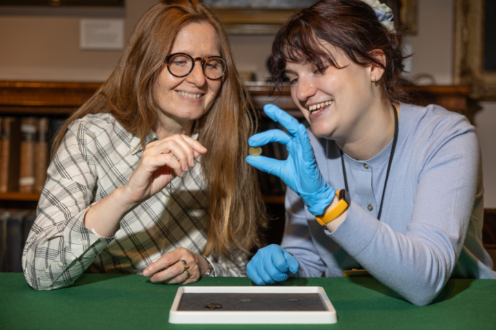 Two women sit at a table smiling. One wears glasses; the other, blue gloves and a blue sweater, holds up a small object, possibly a coin, while the other gestures towards it. A display tray with objects is on the table.