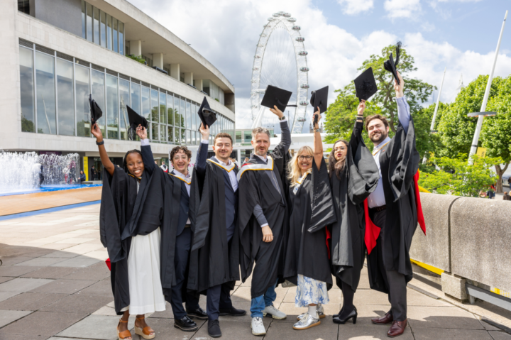 Seven graduates in caps and gowns stand outdoors, smiling and raising their hats in celebration. The London Eye and modern buildings are visible in the background on a sunny day.