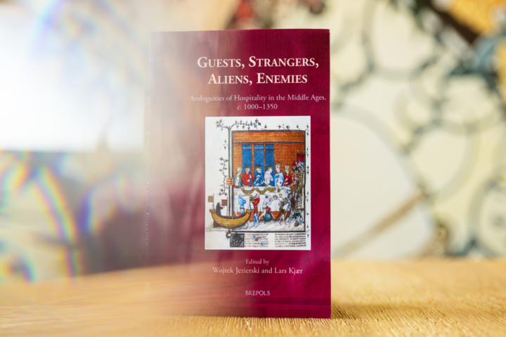 A book titled "Guests, Strangers, Aliens, Enemies" stands upright on a surface. The cover is burgundy with a medieval illustration and white text. The background is softly blurred with abstract patterns.