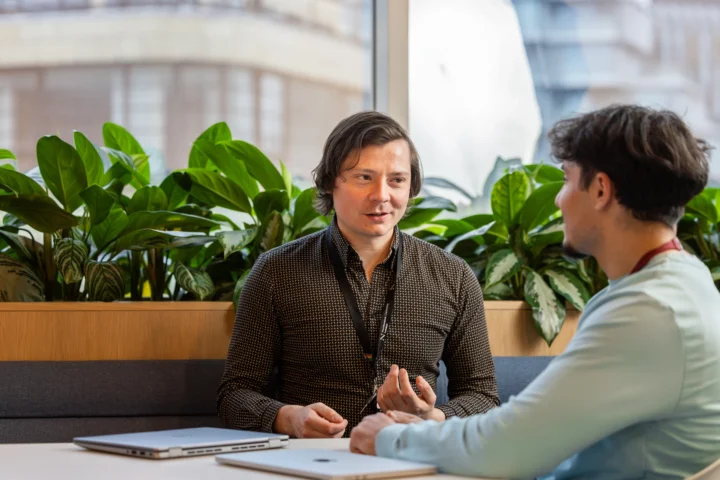 Two men are sitting at a table with laptops in front of them, having a conversation. They are indoors by a window with green plants in the background.