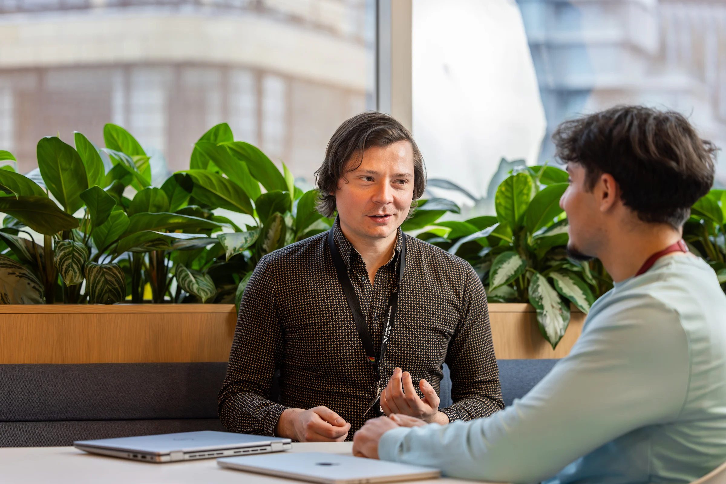 Two men are sitting at a table with laptops in front of them, having a conversation. They are indoors by a window with green plants in the background.
