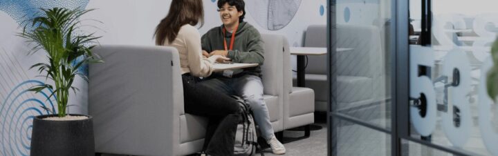 Two people sit across from each other at a small table in a modern office lounge, chatting and smiling. There are gray booths, a potted plant, and glass walls with numbers visible in the foreground.