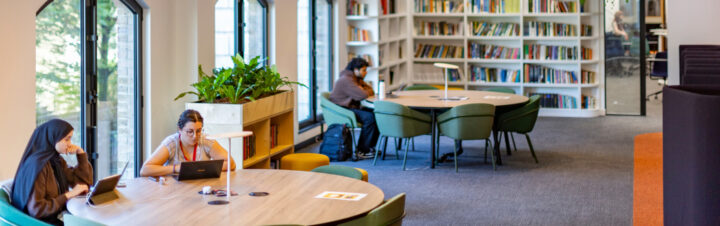 Students study at round tables in a modern library with large windows, bookshelves, and green chairs. Laptops and notebooks are visible on the tables, and potted plants add greenery to the bright, quiet space.