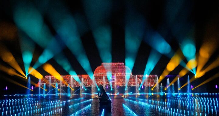A vibrant nighttime light show features colorful beams and patterns projected onto a large building and across water, creating a dramatic and festive atmosphere.