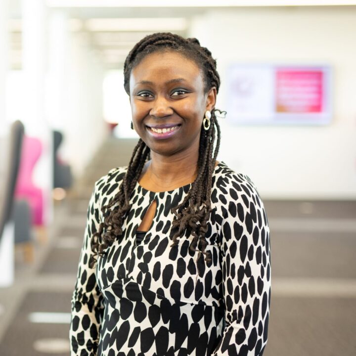 A woman with braided hair, wearing a black and white patterned dress, stands smiling in a bright, modern office space with blurred background.