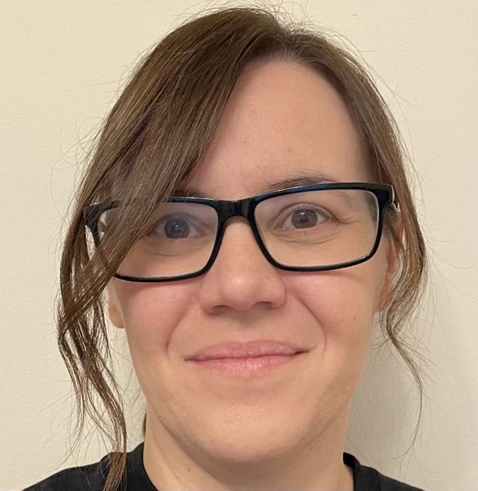 A person with straight brown hair and black-framed glasses smiles softly at the camera. They are standing in front of a plain, light-colored background.