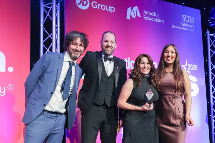 Four people dressed in formal attire smile for a photo at an awards event. One woman holds a glass trophy. The backdrop features logos for JD Group, Mindful Education, and NHS Salisbury.