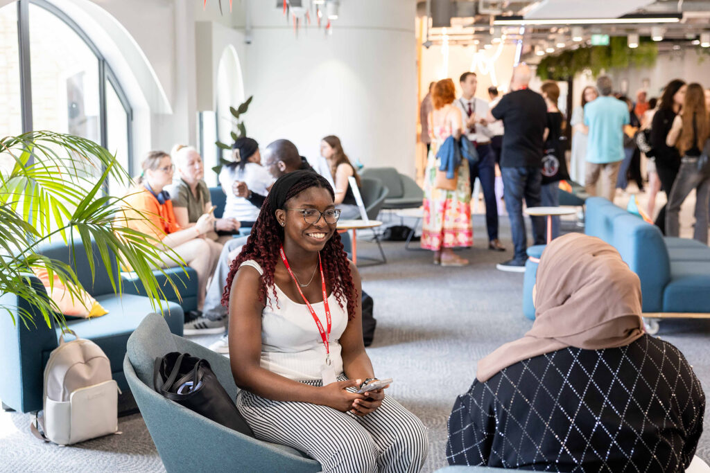 A young woman with glasses and red braids smiles while sitting on a chair, chatting with another person in a busy, modern lounge filled with people talking and relaxing—a welcoming space for student support and connection.