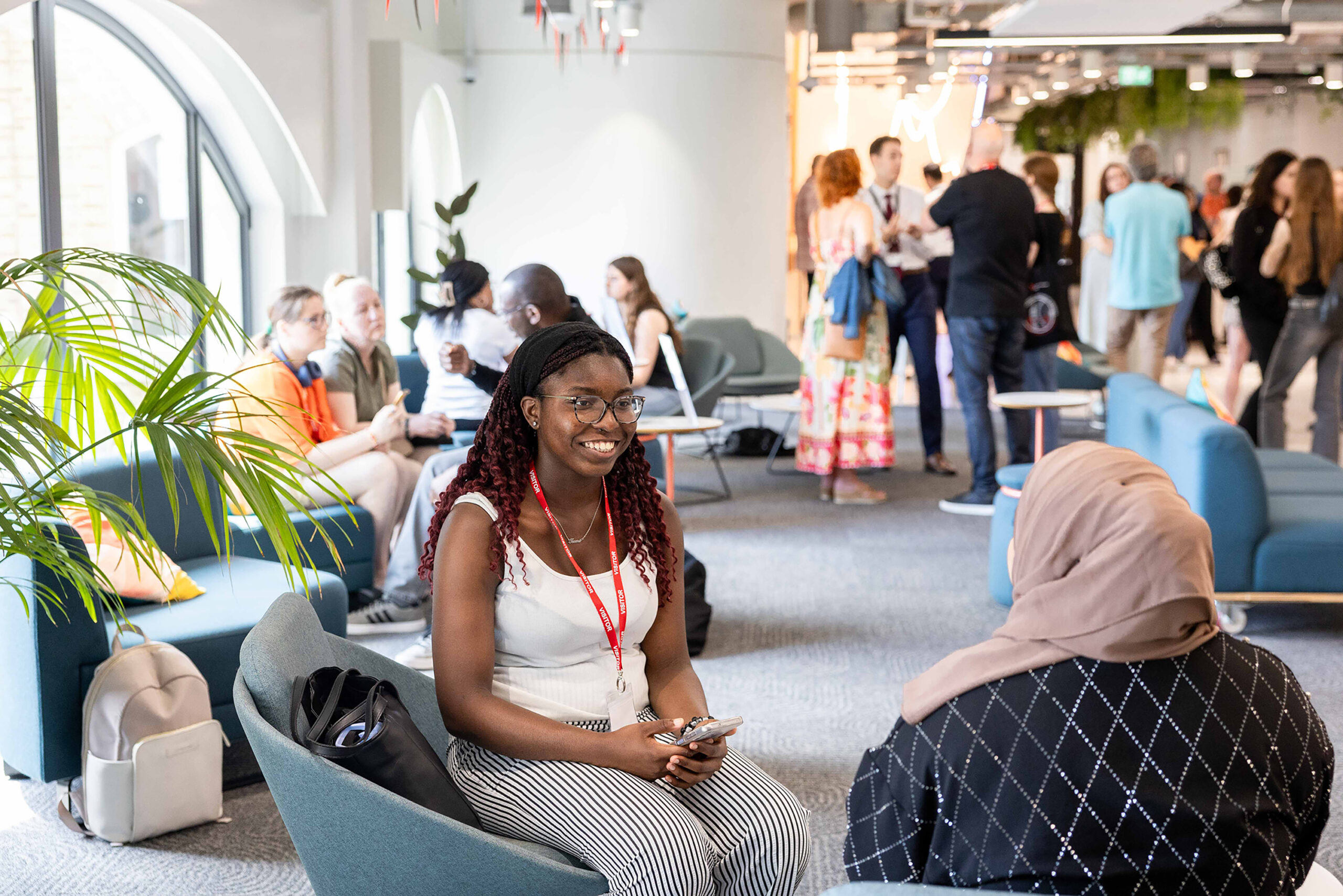 A young woman with glasses and red braids smiles while sitting on a chair, chatting with another person in a busy, modern lounge filled with people talking and relaxing—a welcoming space for student support and connection.