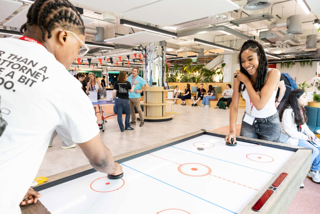 Two young women smile and play air hockey in a bright, modern indoor space with people socializing and watching in the background.