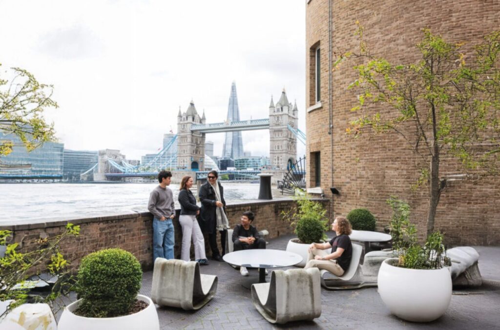Four people stand and sit on a patio with modern white chairs and potted plants, enjoying a clearing view over the River Thames with Tower Bridge and The Shard in the background on a cloudy day.