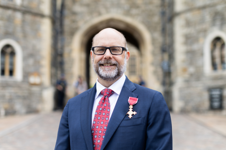 A smiling man with glasses, a gray beard, and a suit with a red patterned tie stands outside a stone building, wearing a medal with a red ribbon pinned to his jacket.