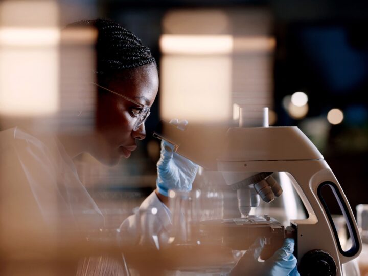 A scientist wearing glasses and blue gloves looks through a microscope in a laboratory, with blurred lights and objects in the foreground.