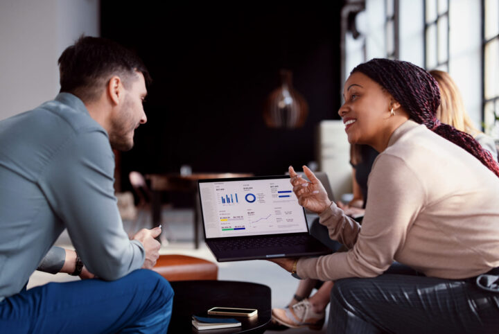 A woman shows a man a laptop screen displaying graphs and charts. They are sitting and discussing in a modern office with other people blurred in the background.
