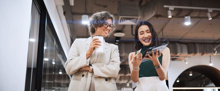 Two women in business attire stand indoors, smiling and laughing together. One holds a coffee cup, while the other shows something on a tablet. The background features modern office lighting and an industrial ceiling.