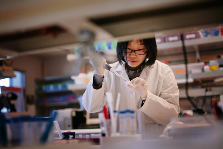 A scientist wearing glasses, gloves, and a lab coat uses a pipette in a laboratory filled with equipment and colorful containers.