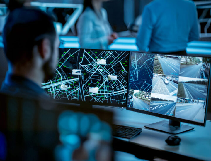 A person monitors multiple computer screens showing a city map with routes and surveillance camera footage of busy roads in a high-tech control room. Other people are blurred in the background.