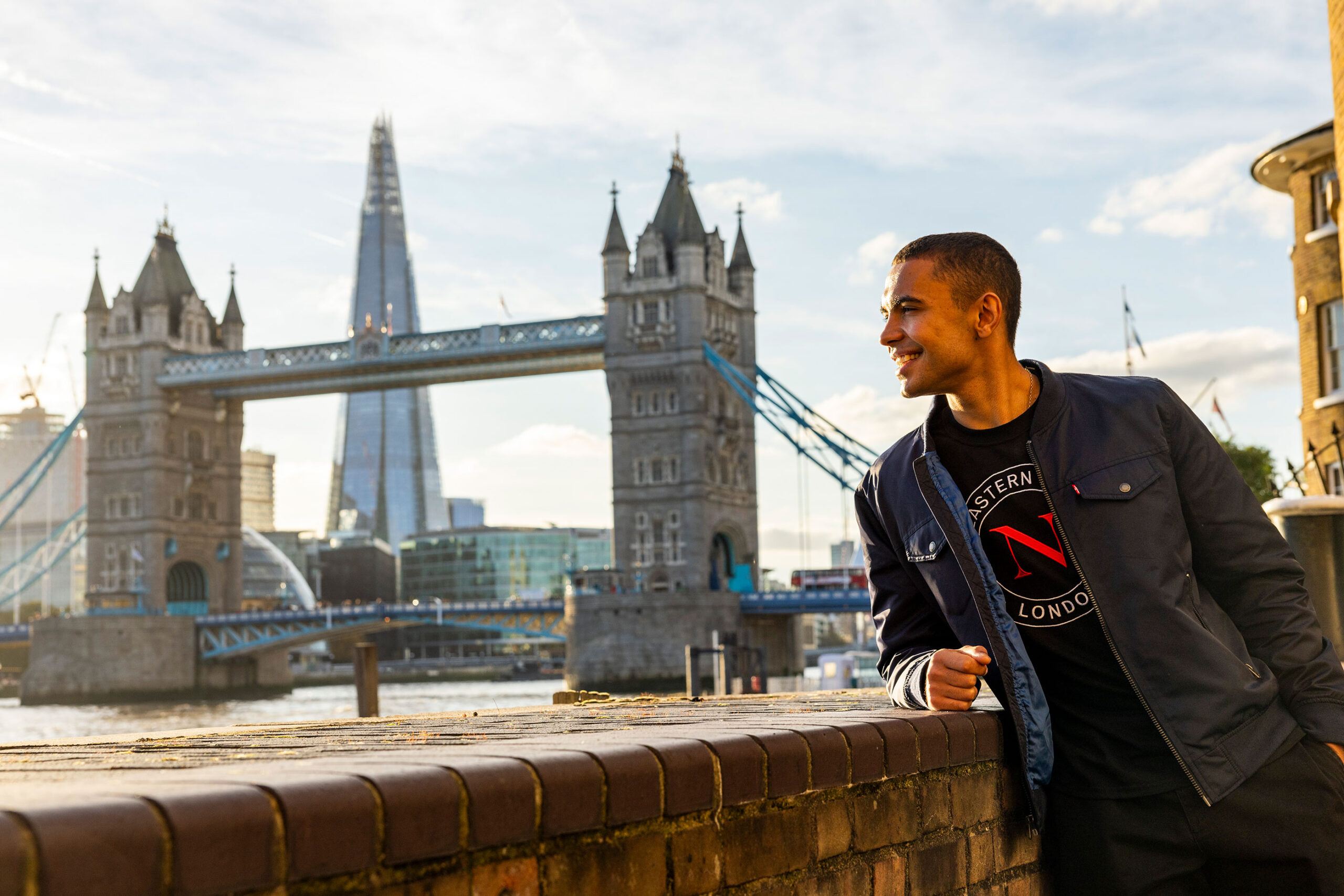 A young man smiles while leaning on a brick wall by the river, with London’s Tower Bridge, The Shard skyscraper, and a nearby university campus in the background on a sunny day.