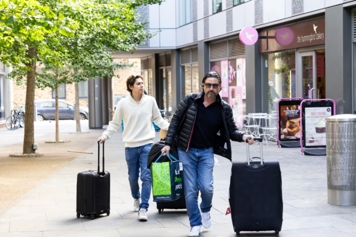Two men walk outside a modern building, each pulling a suitcase. One man wears a black jacket and sunglasses, holding a shopping bag, while the other wears a white sweater. Trees and outdoor seating are visible nearby.