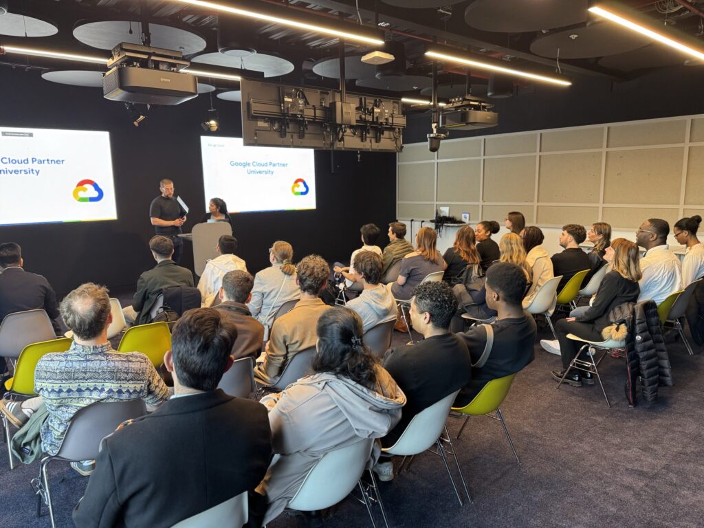 A group of people seated in a modern conference room listen to two presenters at the front, with "Google Cloud Partner University" and "Experiential Trek" displayed on screens behind them.