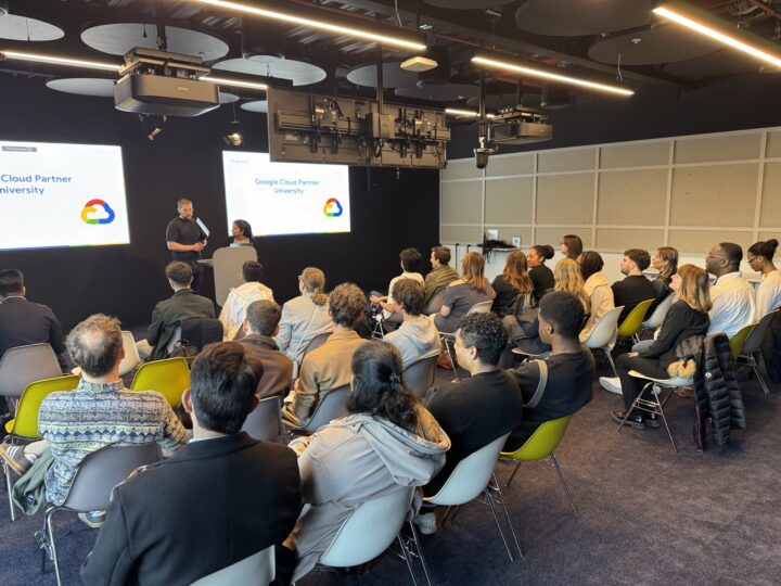 A group of people seated in a modern conference room listen to two presenters at the front, with "Google Cloud Partner University" and "Experiential Trek" displayed on screens behind them.