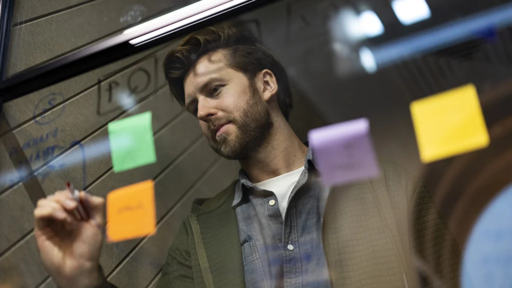 A man, likely pursuing masters-degrees, writes on a transparent board covered with colorful sticky notes, appearing focused and thoughtful in a modern, indoor setting.