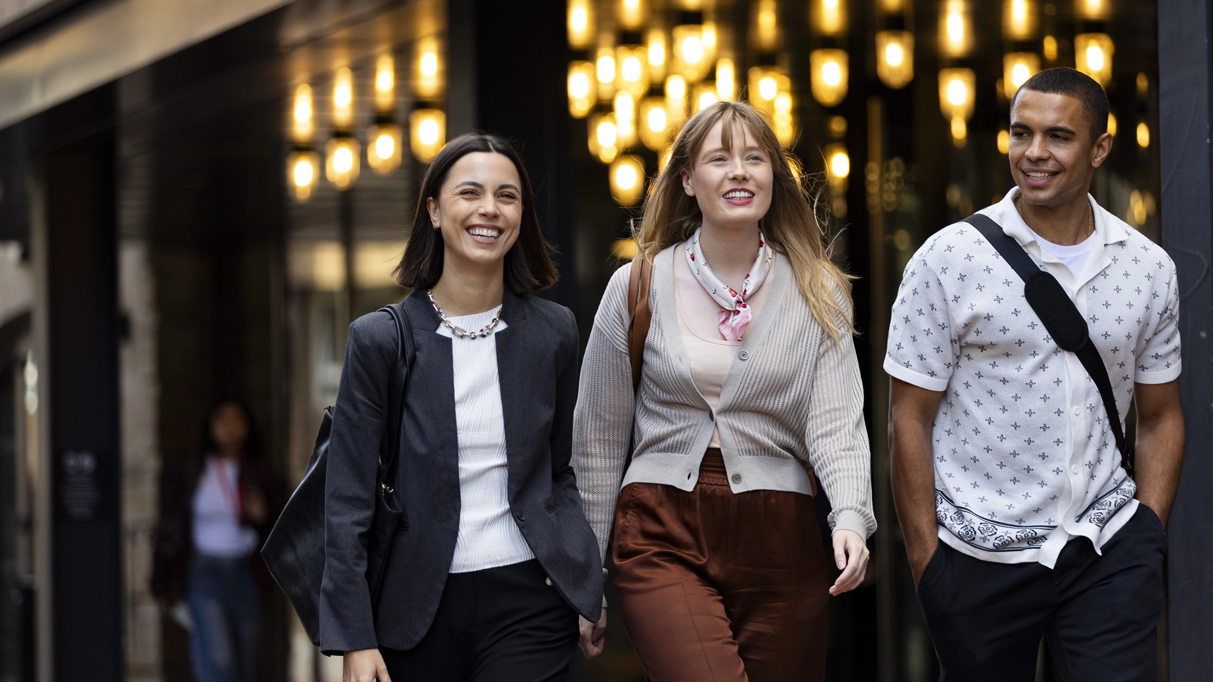 Three young adults, two women and one man, walk together outdoors near a university campus, smiling and talking. They are dressed in smart casual clothing, with warm hanging lights illuminating the background.