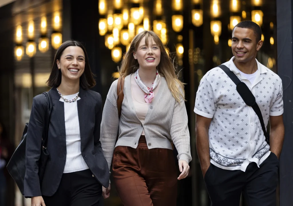 Three young adults, possibly pursuing masters-degrees, walk together outside a building with hanging lights, smiling and engaged in conversation. They wear casual and business-casual clothing.