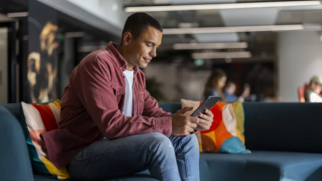 A man sits on a blue couch in a modern office, looking at a tablet. He wears a red shirt and jeans, with colorful pillows beside him. The background is blurred, showing other people and office lighting.