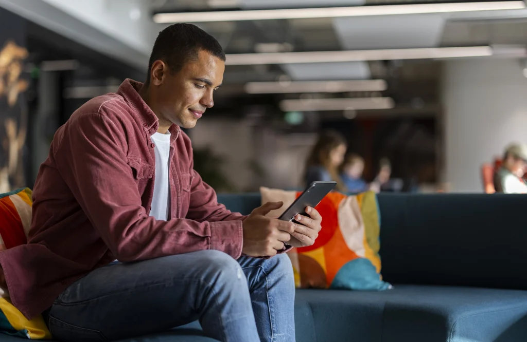 A man sits on a blue couch in a modern office, looking at a tablet. He wears a red shirt and jeans, with colorful pillows beside him. The background is blurred, showing other people and office lighting.