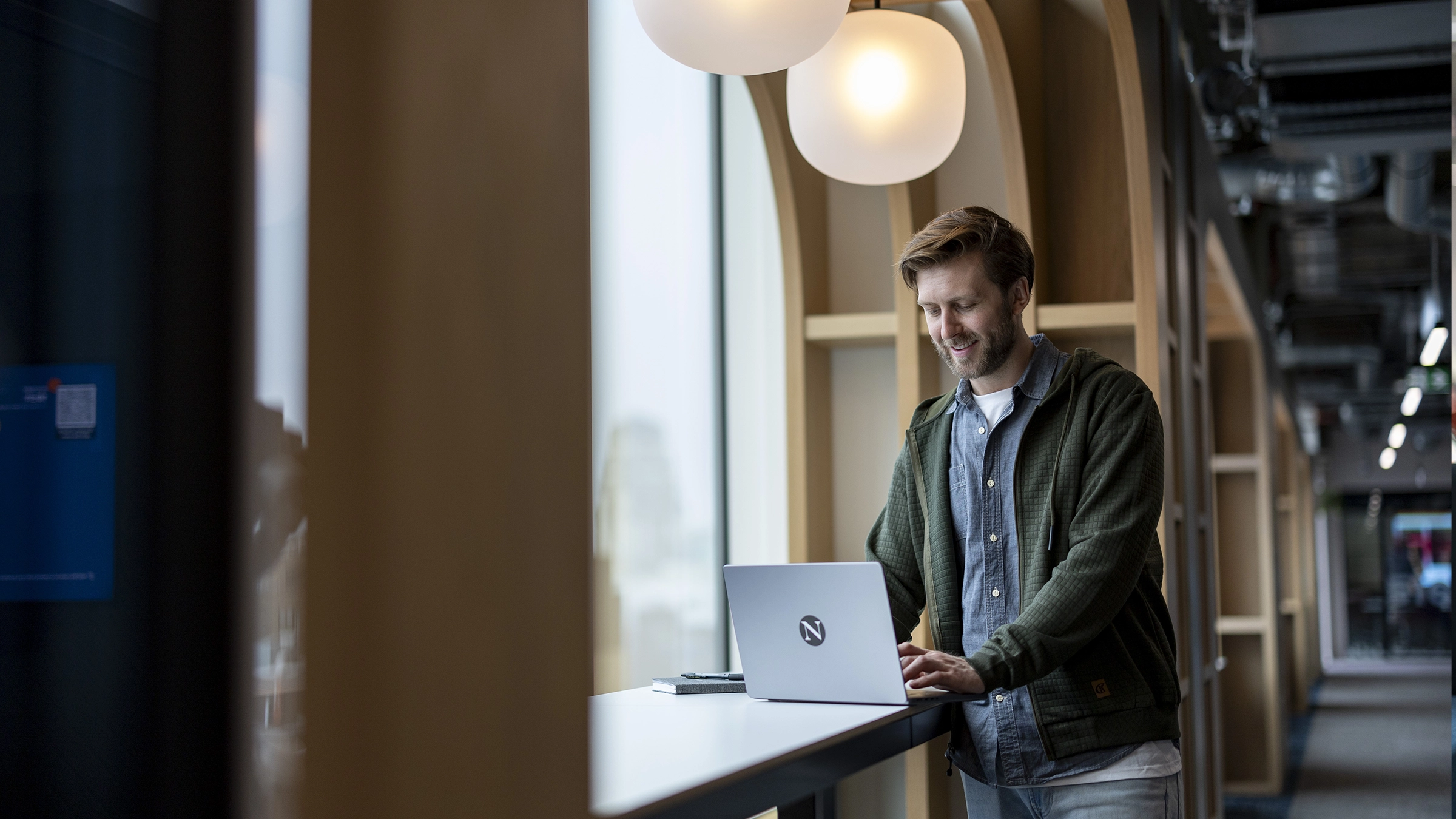A man stands at a high desk by large windows in a modern office, smiling while working on a laptop. The bright workspace, illuminated by hanging lights and wooden arch details, reflects the inspiring atmosphere ideal for pursuing masters-degrees.