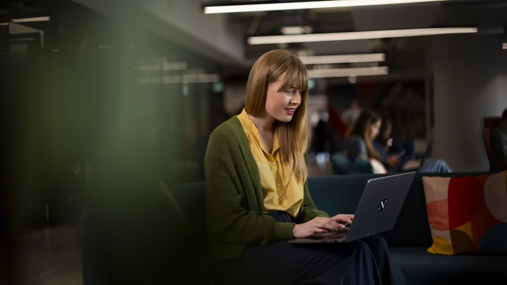 A woman with long blonde hair, wearing a yellow blouse and green cardigan, sits on a dark sofa researching Masters Tuition Fees on her laptop in a modern, well-lit office space while others work in the background.