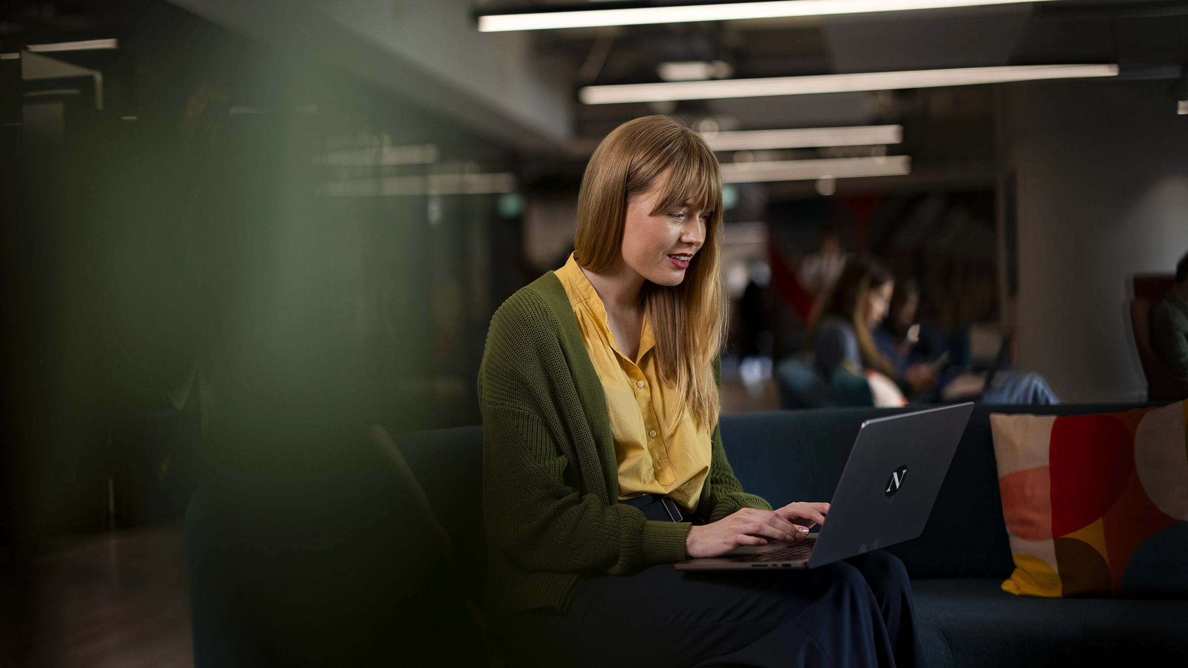 A woman with long blonde hair, wearing a yellow blouse and green cardigan, sits on a dark sofa researching Masters Tuition Fees on her laptop in a modern, well-lit office space while others work in the background.