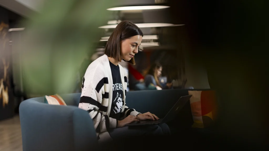 A woman with a bob haircut sits on a blue couch using a laptop, researching Master’s tuition fees. She wears a striped cardigan and black shirt, concentrating on her work. The background is softly blurred, with another person visible.
