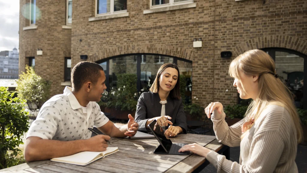 Three people sit at a wooden table outside, having a discussion about masters-degrees. One woman gestures towards a tablet, while the other woman and a man with a notebook listen. A brick building is in the background.