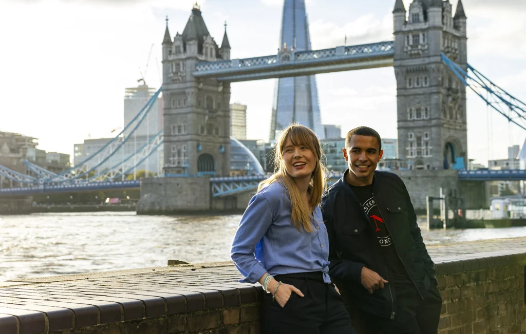 Two people smiling and posing by a brick wall along the River Thames, with Tower Bridge and the Shard skyscraper in the background—a classic London scene popular with students pursuing masters-degrees.