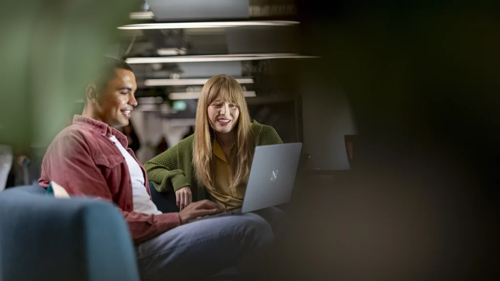 Two people sit together, smiling and looking at a laptop screen in a modern, dimly-lit indoor setting. The relaxed atmosphere suggests they might be exploring masters-degrees or collaborating on further education plans.