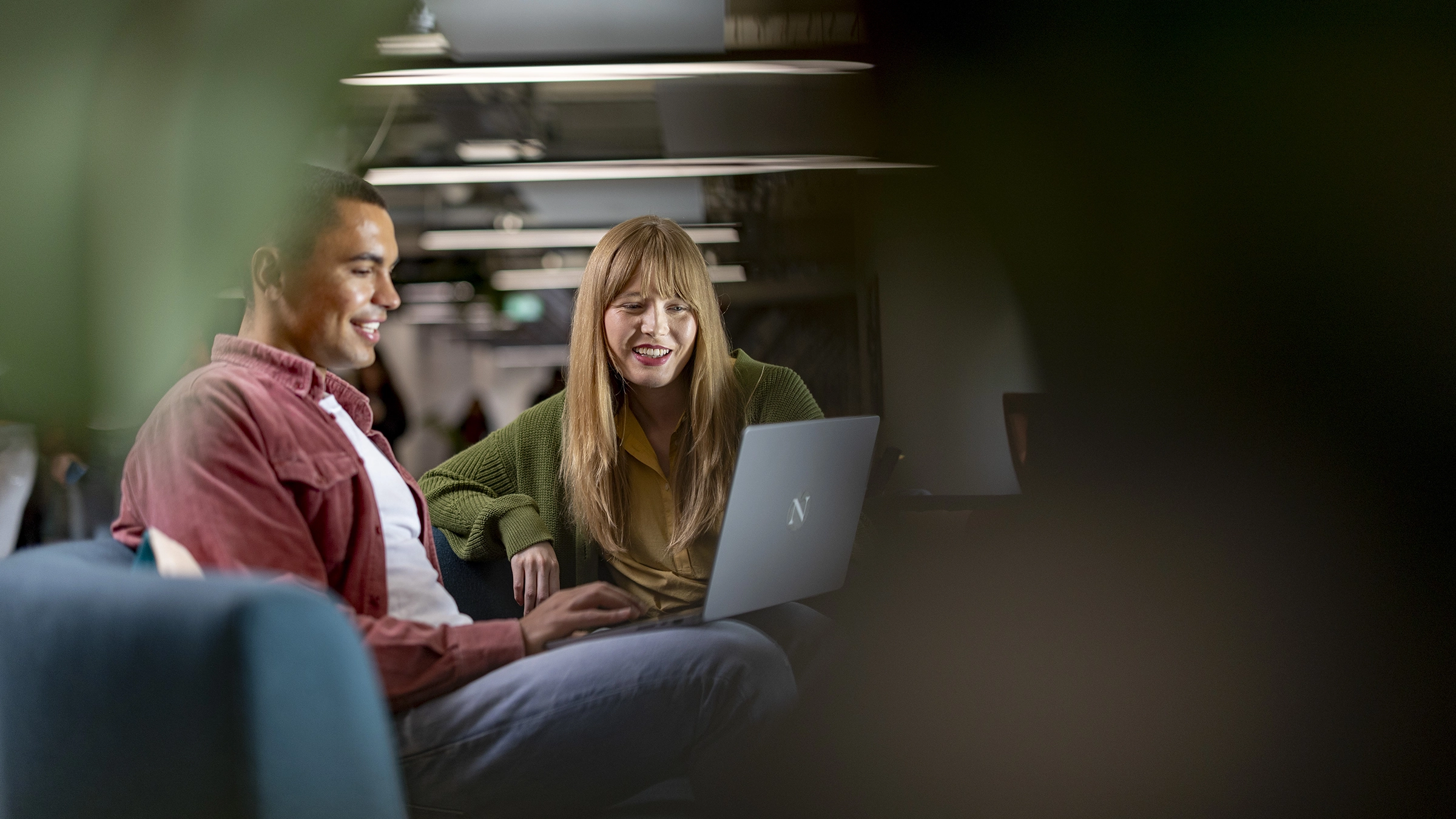 Two people sit together, smiling and looking at a laptop screen in a modern, dimly-lit indoor setting. The relaxed atmosphere suggests they might be exploring masters-degrees or collaborating on further education plans.