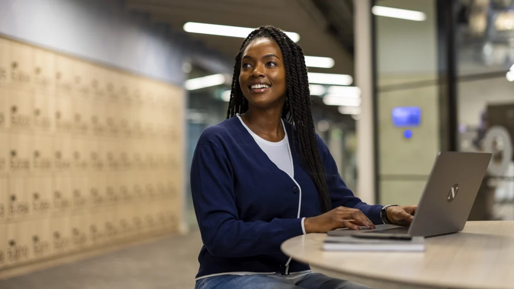 A woman with braided hair, wearing a navy sweater and jeans, sits at a round table in a modern hallway, smiling while exploring masters-degrees on her laptop. Lockers and soft lighting are visible in the background.