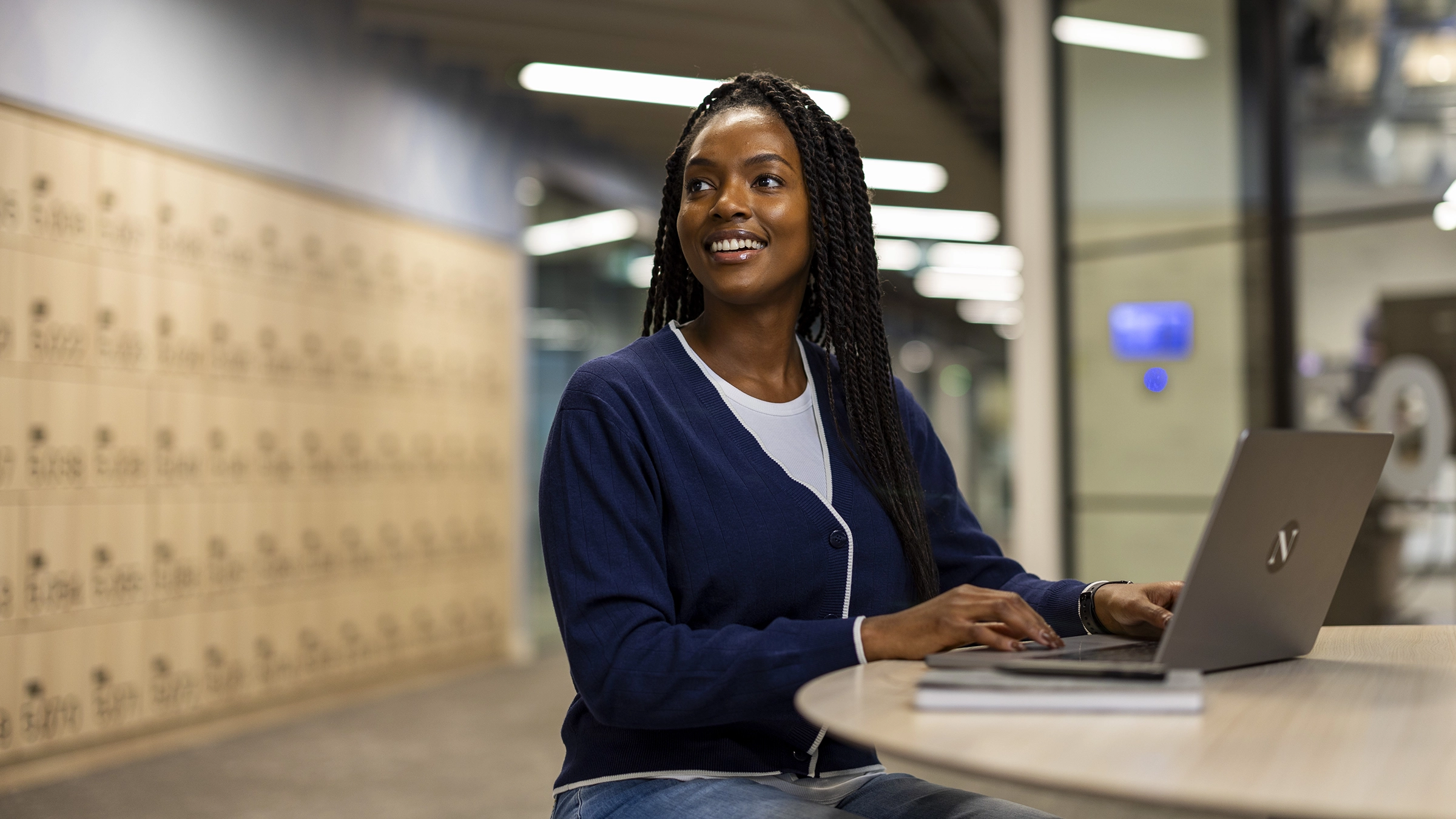 A woman with braided hair, wearing a navy sweater and jeans, sits at a round table in a modern hallway, smiling while exploring masters-degrees on her laptop. Lockers and soft lighting are visible in the background.