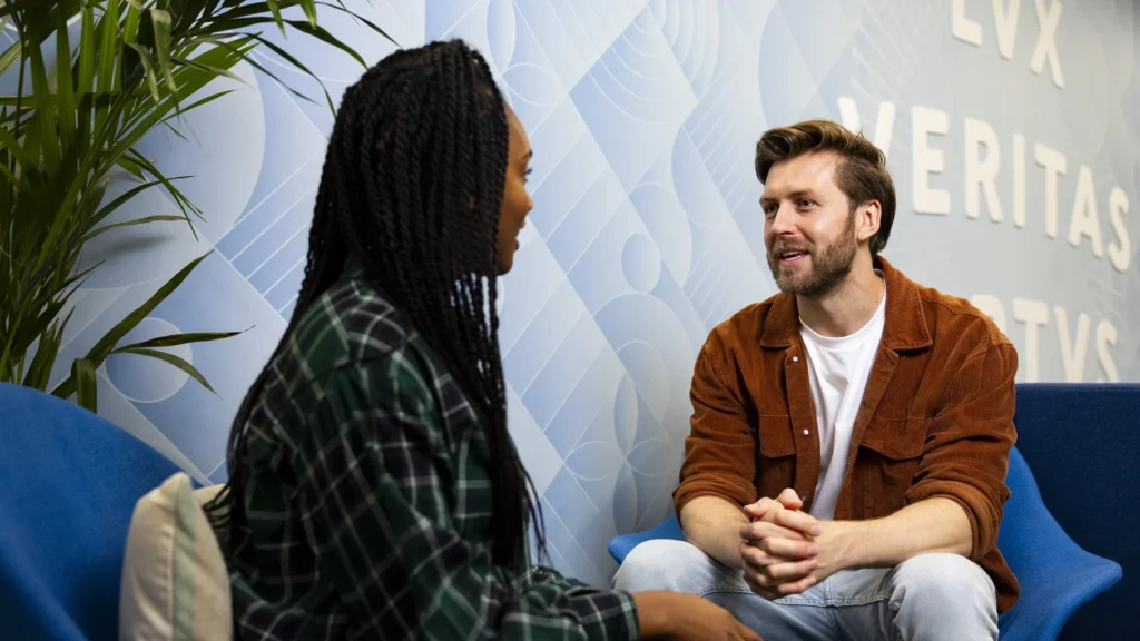 Two commuter students sit in modern blue chairs, having a conversation. The person on the left has braided hair and a green plaid shirt, while the one on the right has light brown hair, a beard, and a brown jacket. A plant and blue wall are in the background.
