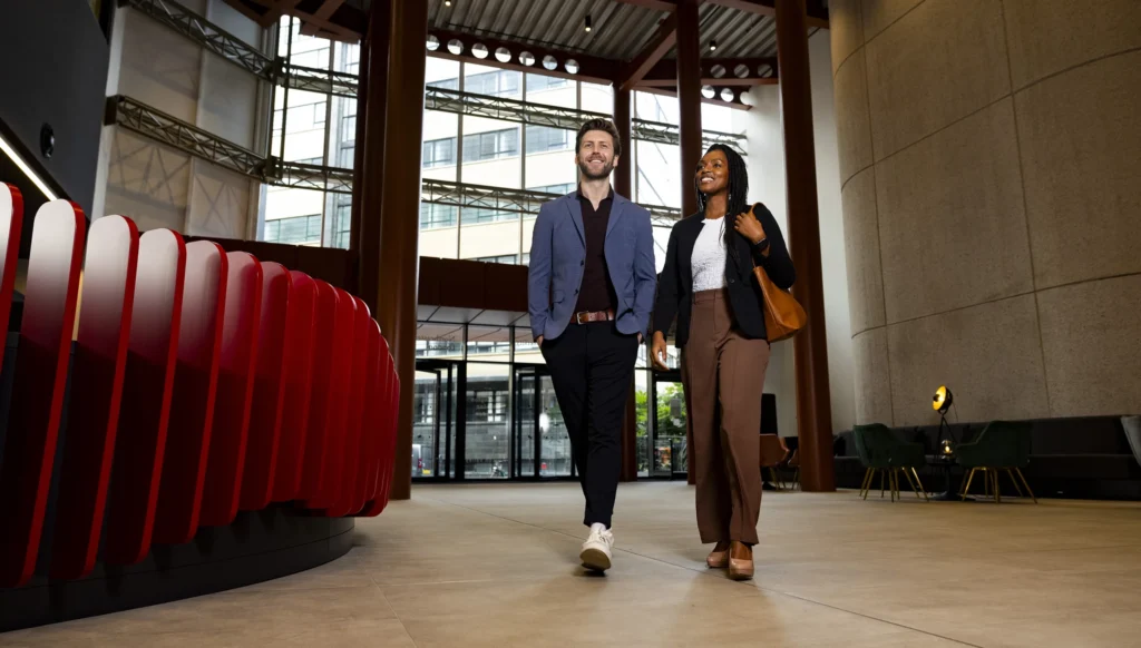 A man and a woman in business attire, both discussing their masters-degrees, walk together through a modern, spacious office lobby with large windows, high ceilings, and contemporary furniture.
