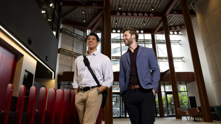 Two men in business attire walk and smile together in a modern, open office lobby with high ceilings and red architectural accents, discussing their recent achievements earning masters-degrees.