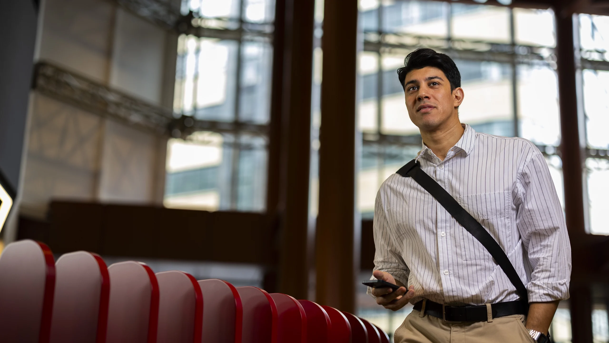 A man in a striped shirt with a shoulder bag stands indoors, holding a smartphone. He looks ahead thoughtfully, as if considering masters-degrees, with modern glass windows and architectural features in the background.