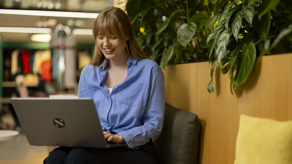 A woman with light brown hair and a blue shirt sits on a cushioned bench, smiling while using her laptop to research Master's Admissions. She is indoors, next to a large green plant and a wooden partition.